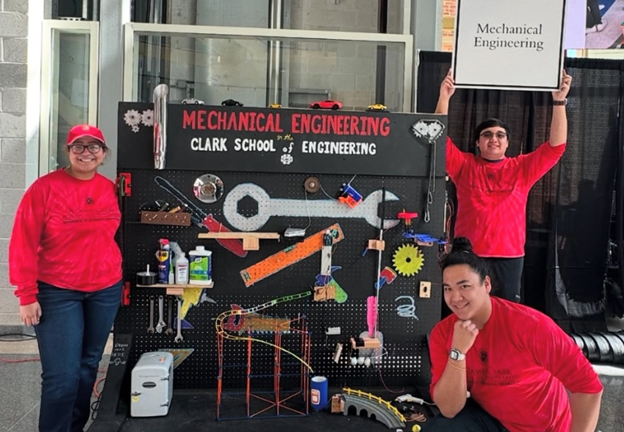 Students in front of a mechanical engineering sign with a wrench and gadgets. One student holds a UMD Mechanical Engineering sign