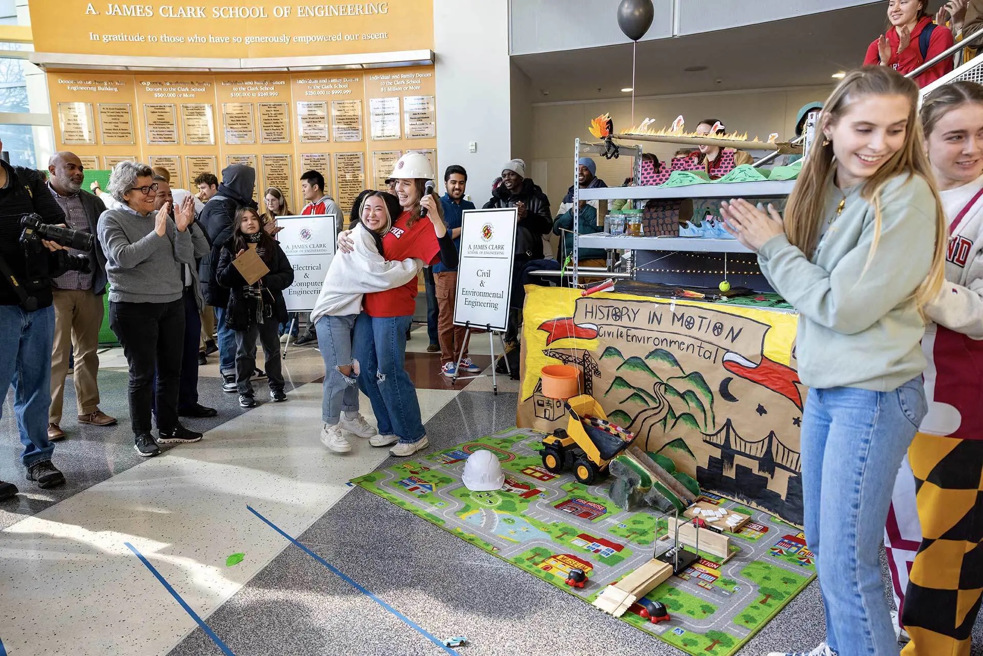 Civil engineering students Genevieve Sullivan ’25 (center, hat) and Mai Brosseau ’25 embrace after their team’s Rube Goldberg machine—which celebrates the discipline’s history and achievements—successfully launched the toy car.