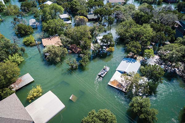 Aerial photo showing a flooded area with trees, the tops of houses, and a power line above the water line. A small group of people on a boat can be seen in the middle.