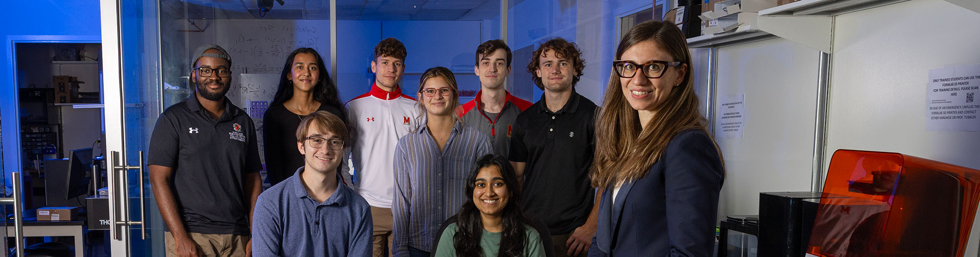 A professor sits in a robotics lab with a group of students next to her