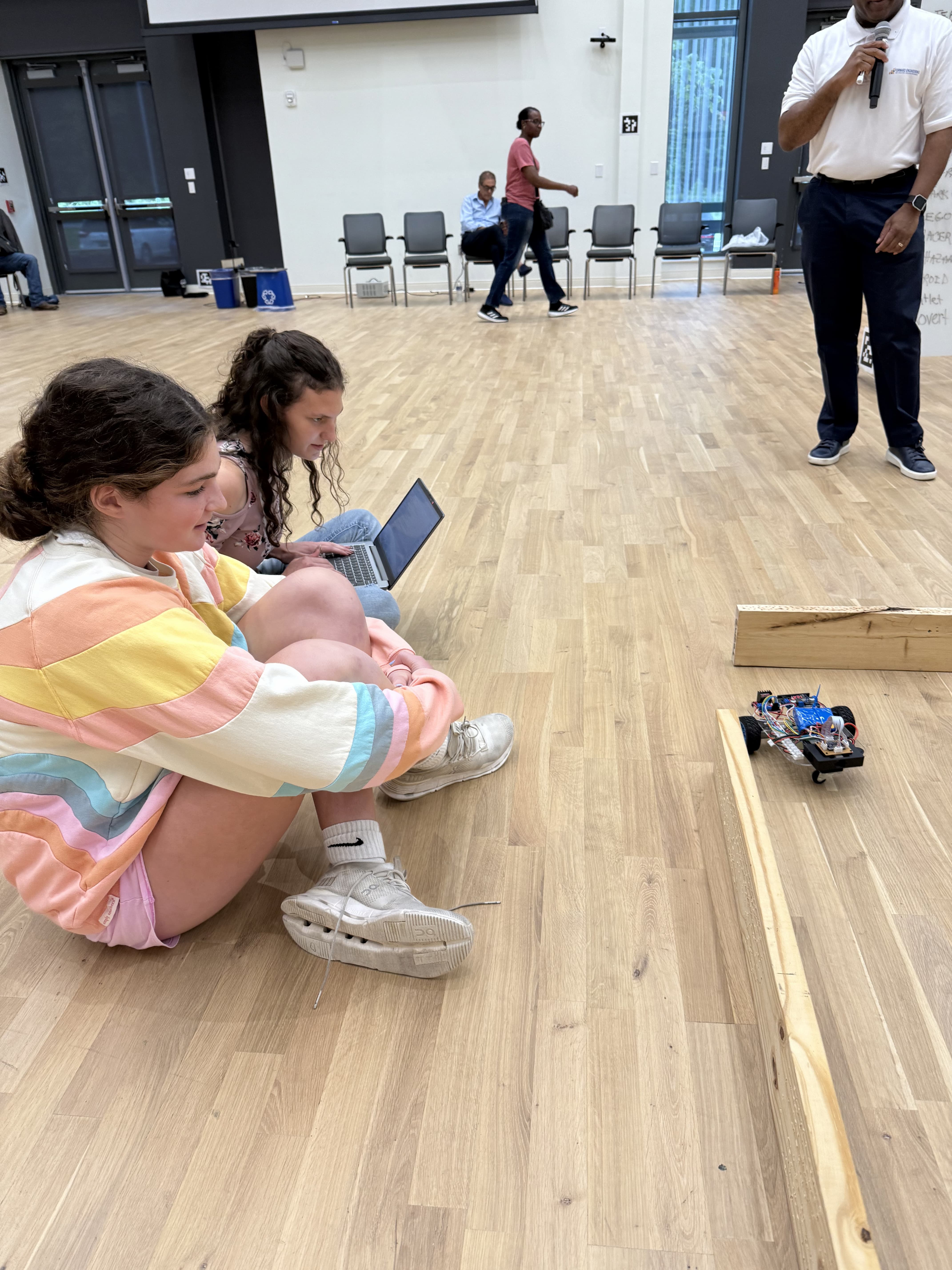 student working on a laptop to control a robot through a maze while classmate looks on.
