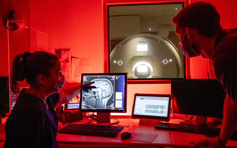 two students with masks on work in a room with red light in front of a computer screen with an MRI image of a human head and brain