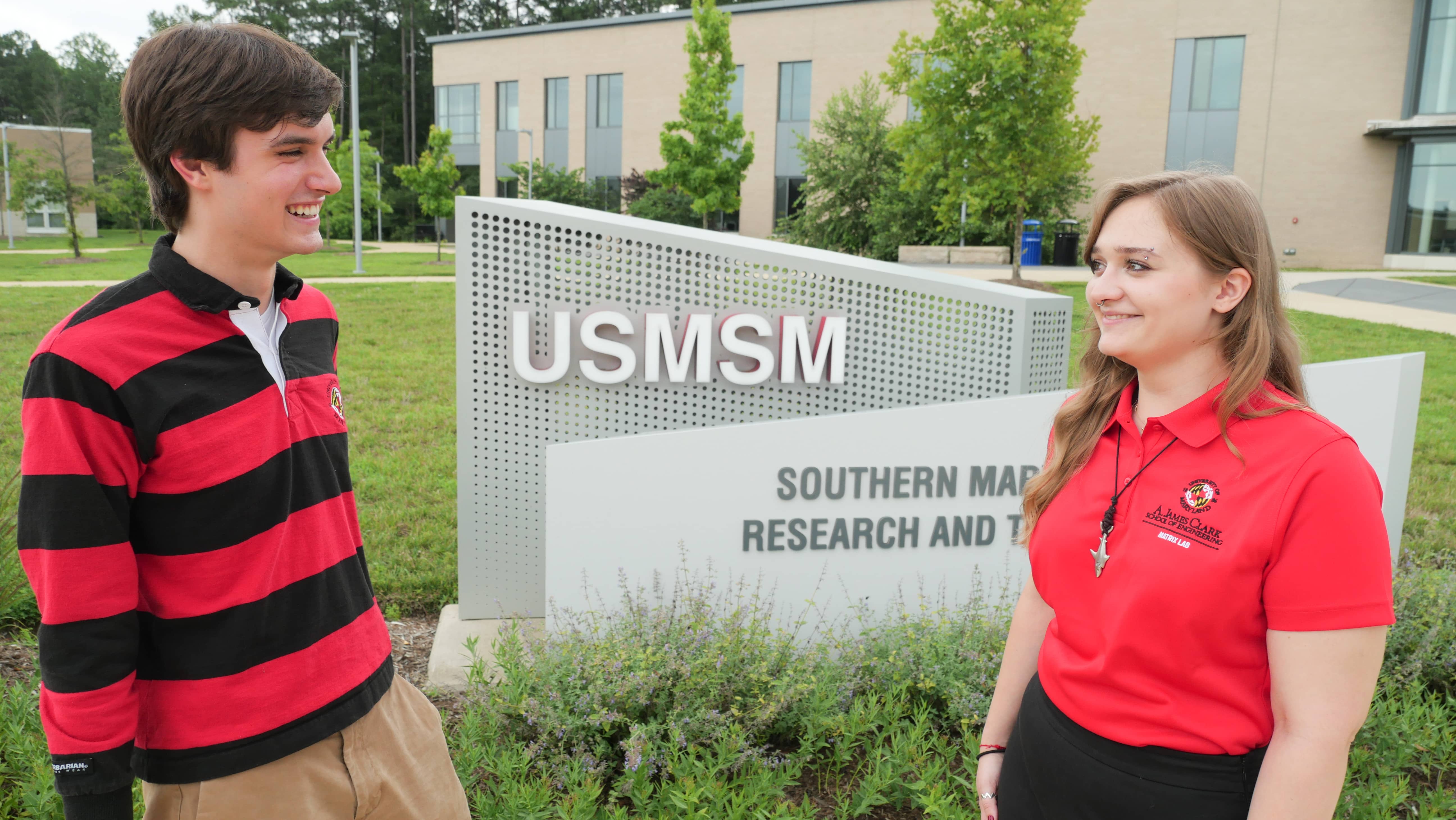 Students smiling while facing each other in front of USMSMS sign while wearing Clark School gear