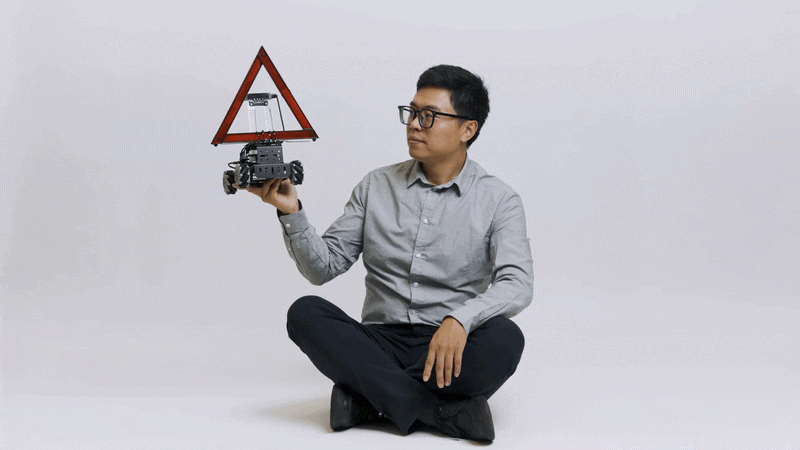 Terry Yang sits on the floor and holds up a small autonomous vehicle with a large reflective triangle