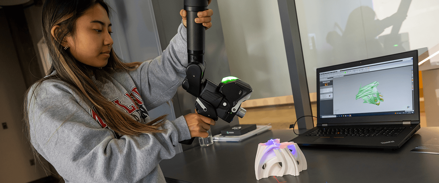 a student in a gray sweatshirt uses a robotic arm to finalize her 3D design with a computer on the table in the background