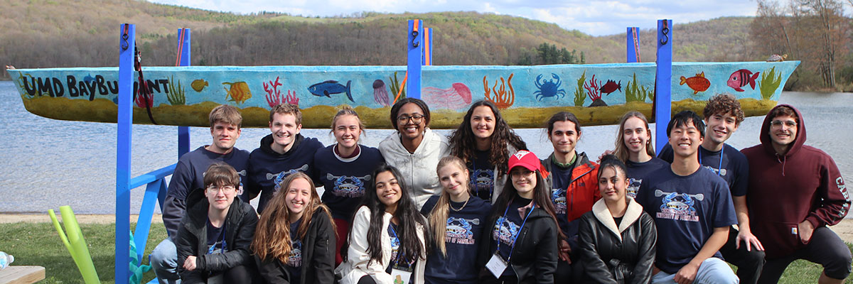 Sixteen students pose in front of a brightly colored canoe by the Bay