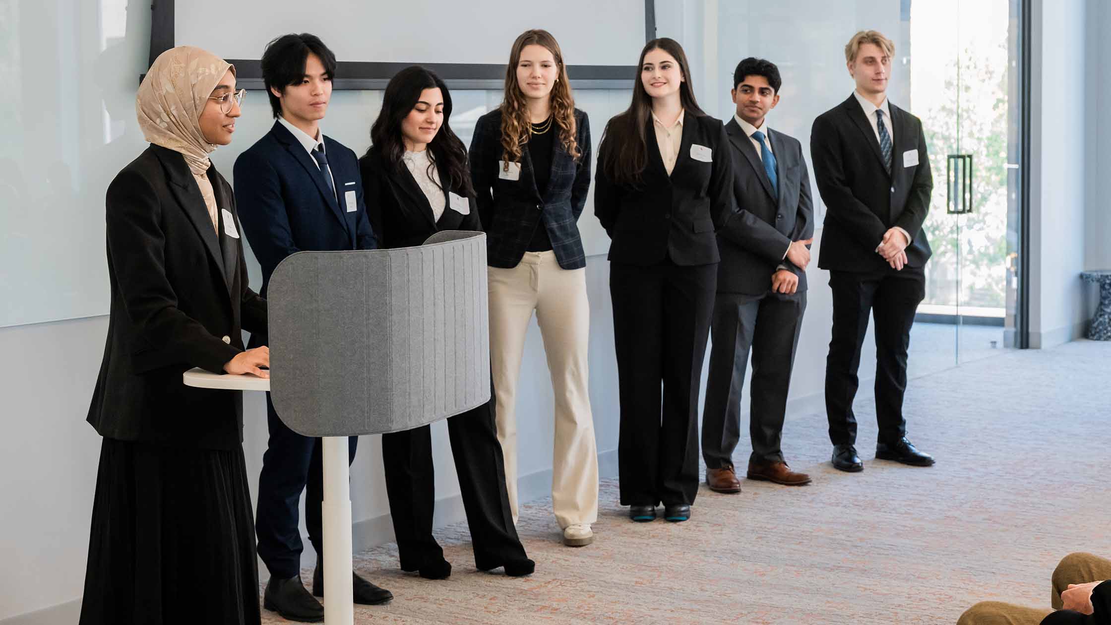 Seven students in suits stand at the front of a room. One speaks from a lectern