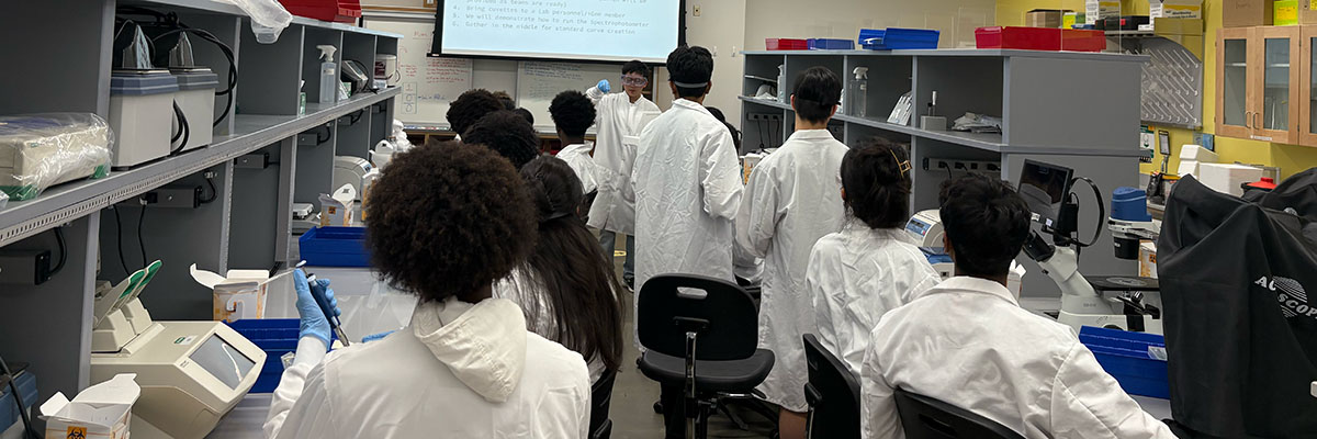 12 students in lab coats watch another student in a lab coat hold up a vial