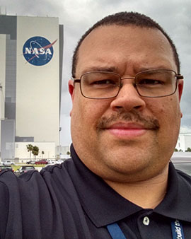 selfie of Marcellus Proctor wearing glasses and a black polo shirt standing next to a building branded with a large blue NASA logo