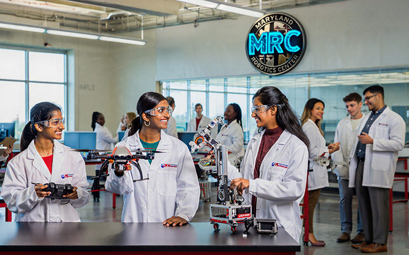 Students wearing lab coats and safety glasses hold robots under a sign reading Maryland Robotics Center
