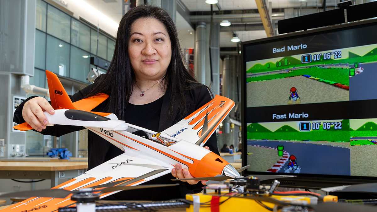 Professor Mumu Xu holds a model plane. A screen with a Mario Kart test is behind her and a drone in front of her.