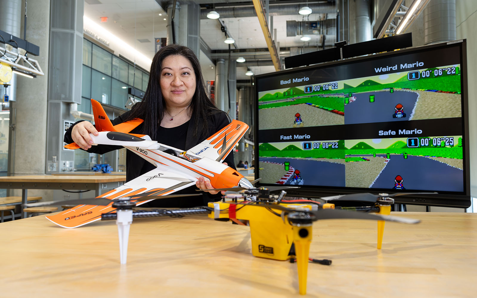 Professor Mumu Xu holds a model plane. A screen with a Mario Kart test is behind her and a drone in front of her.