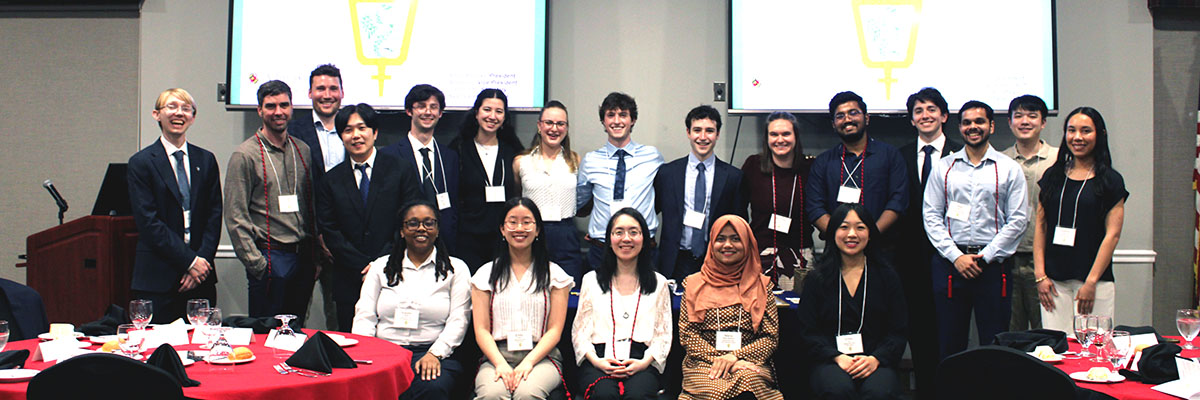 20 students pose with smiles in front of two projector screens