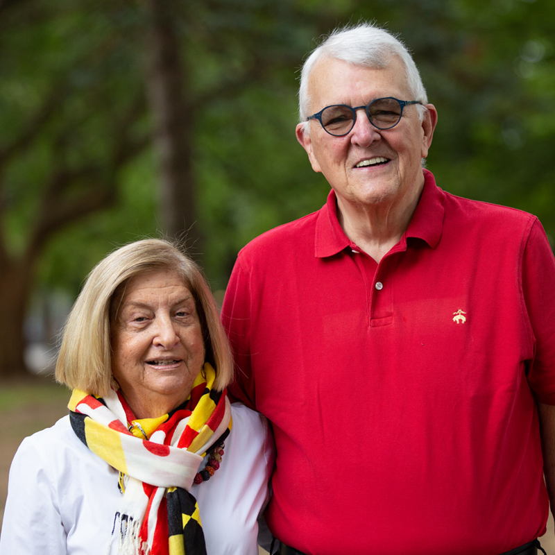 A woman wearing a patterned scarf in Maryland colors and a white shirt stands next to a man in a red polo. They are smiling at the camera.