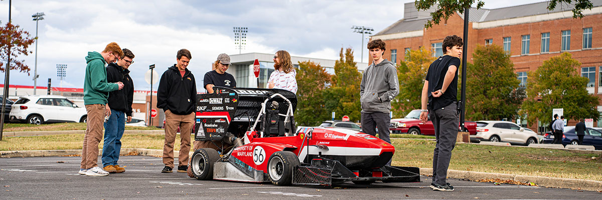 Seven students stand near a bright red formula-style race car parked outside on the UMD campus