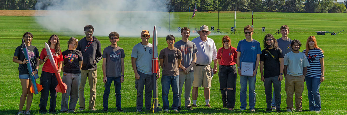 15 people (students and advisors) stand outside on a field, many holding small rockets