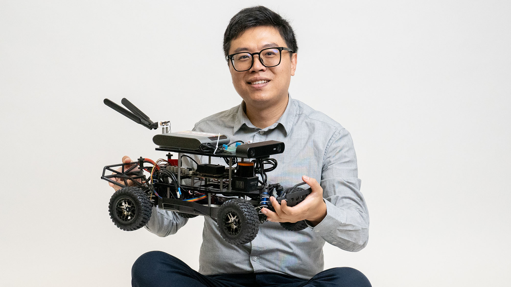 Terry Yang sits on the floor and holds up a small autonomous vehicle with a large reflective triangle