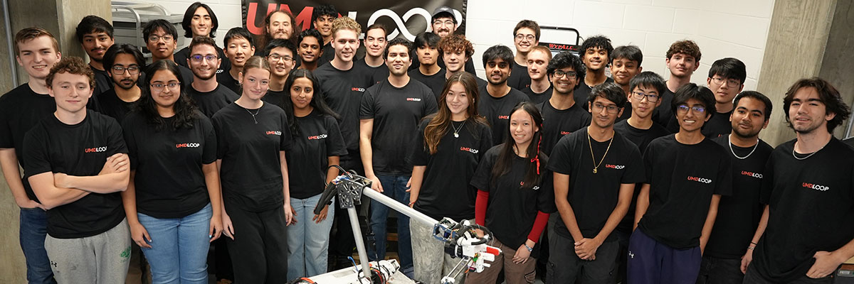 A large group of people wearing black T-shirts stand behind a rocket with a UMD Loop banner behind them